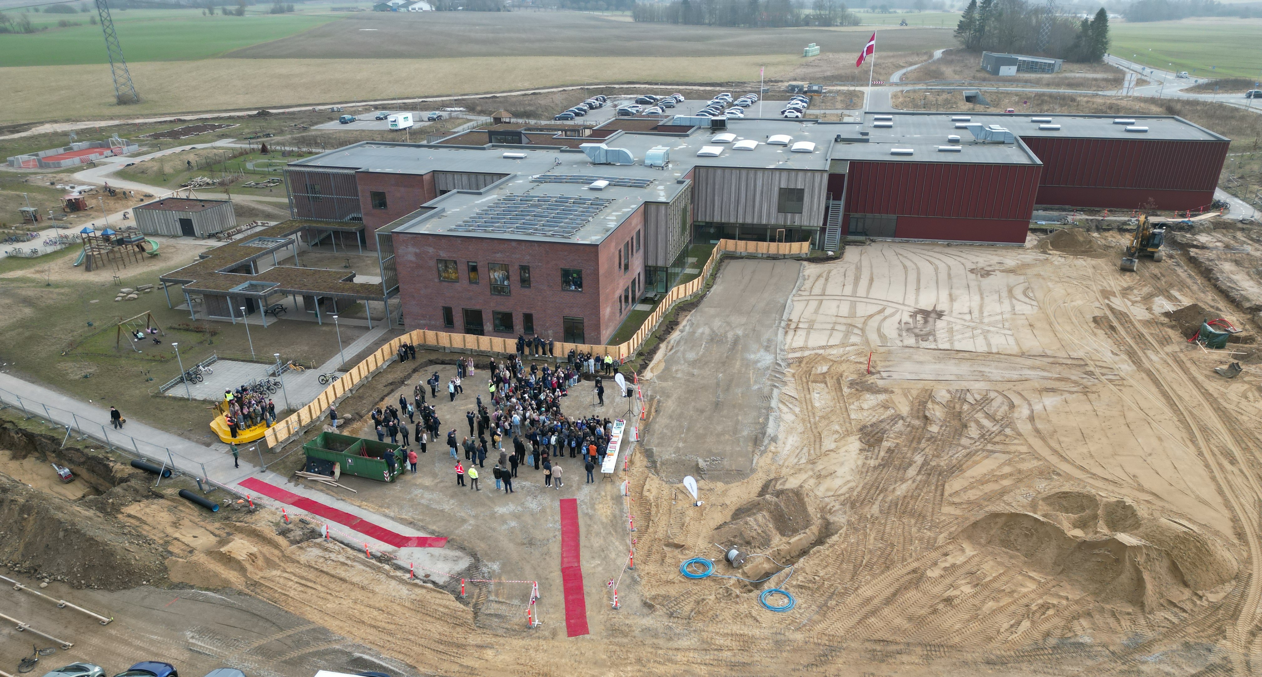 Dronebillede af Sommerbæk Skole og det område, hvorpå det nye byggeri skal ske. Billedet er taget på sagen for spadestikket og en gruppe mennesker ses. Foto: Skanderborg Kommune